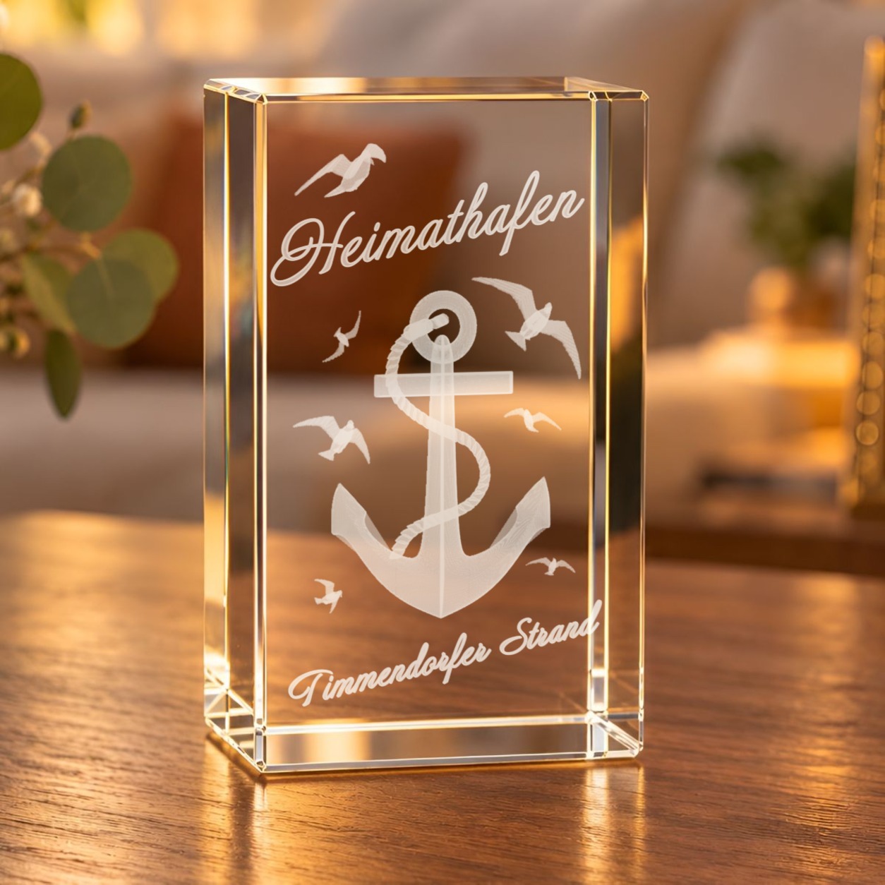 3D-Glaskristall Anker mit Möwen und Schriftzug „Heimathafen Timmendorfer Strand“ auf einem Holztisch im Wohnraum